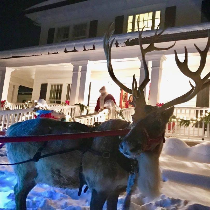 A reindeer stands by a house and sleigh in a winter landscape.